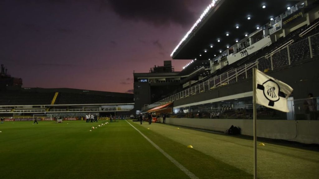 Vista do gramado e das arquibancadas do Estádio Vila Belmiro, em Santos, com céu claro ao fundo.