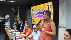 Secretária municipal discursa ao microfone durante o lançamento oficial do Carnaval 2026 de Campinas, em evento com autoridades sentadas à mesa e telão com identidade visual da festa ao fundo