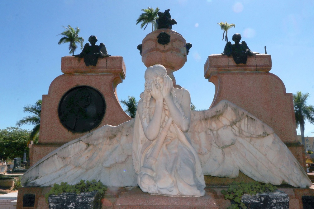 Escultura de anjo com asas abertas e rosto coberto pelas mãos em túmulo do Cemitério da Saudade, em Campinas, com palmeiras ao fundo.