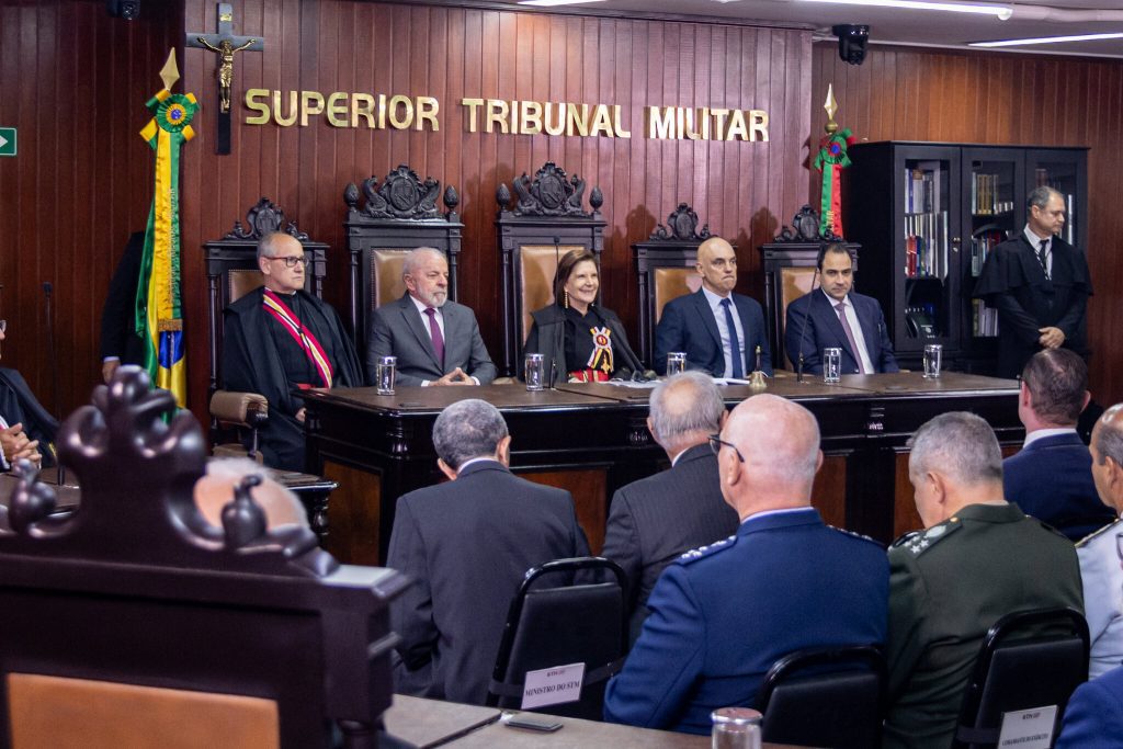 Plenário do Superior Tribunal Militar (STM) com ministros sentados em bancadas semicirculares de madeira escura. Ao fundo, destaca-se o brasão da República e as bandeiras do Brasil.