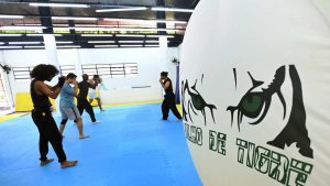Alunos praticam artes marciais em aula no Espaço Arte e Movimento, no Centro de Campinas, em sala com tatame azul e parede com ilustração de olhos e a inscrição “Olho de Tigre”.