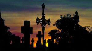 Cruz e túmulos em silhueta no Cemitério da Saudade, em Campinas, ao pôr do sol, com céu alaranjado ao fundo.