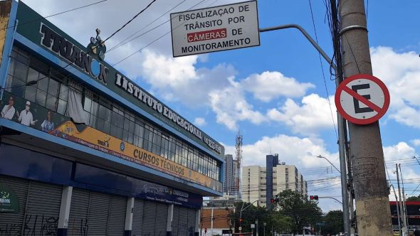 Placa indica fiscalização de trânsito por câmeras em cruzamento da Avenida João Jorge, em Campinas, com sinal de proibido estacionar e semáforo ao fundo.