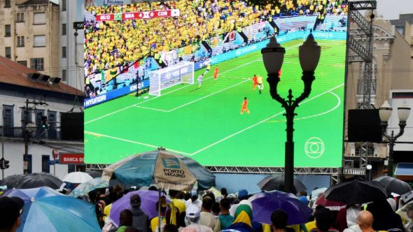 Público acompanha transmissão de jogo da Seleção Brasileira em telão durante evento ao ar livre em área urbana.