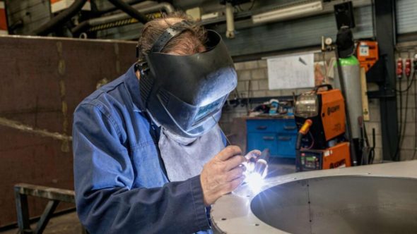 Trabalhador realiza soldagem em peça metálica dentro de ambiente industrial, utilizando máscara de proteção e equipamentos de segurança.